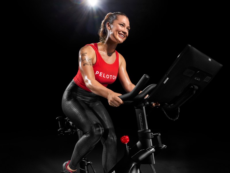 Frau lachend auf schwarzem Peloton Bike mit rotem Peloton-Shirt vor dunklem Hintergrund