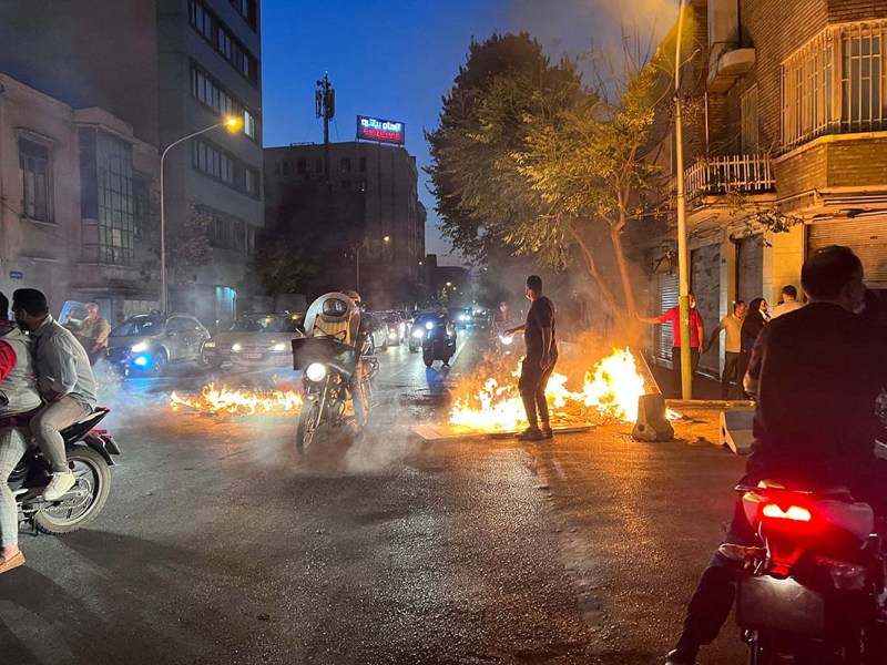 Rauch und Feuer erhellen die Dämmerung einer Straßenszene. Ein Mensch ist als Silhouette zu erkennen.