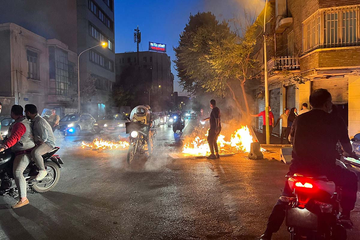 Rauch und Feuer erhellen die Dämmerung einer Straßenszene. Ein Mensch ist als Silhouette zu erkennen.