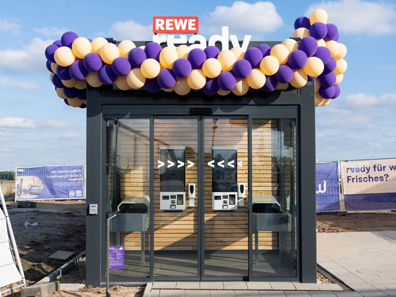 Rewe ready besteht aus einem holzverkleideten Lager und einem kleinen, verglasten Eingangsbereich mit je zwei Tochscreens und Laufbändern. Zur Eröffnung war die Mini-Filiale mit gelben und lilafarbenen Luftballons dekoriert.