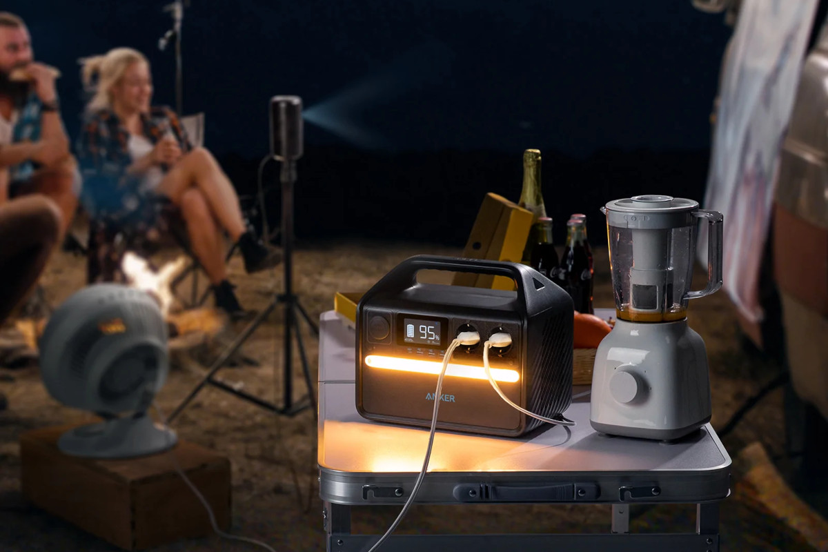 Leuchtende Power Station von Anker auf Campingtisch neben Mixer, im Hintergrund Mann und Frau in Campingstühlen