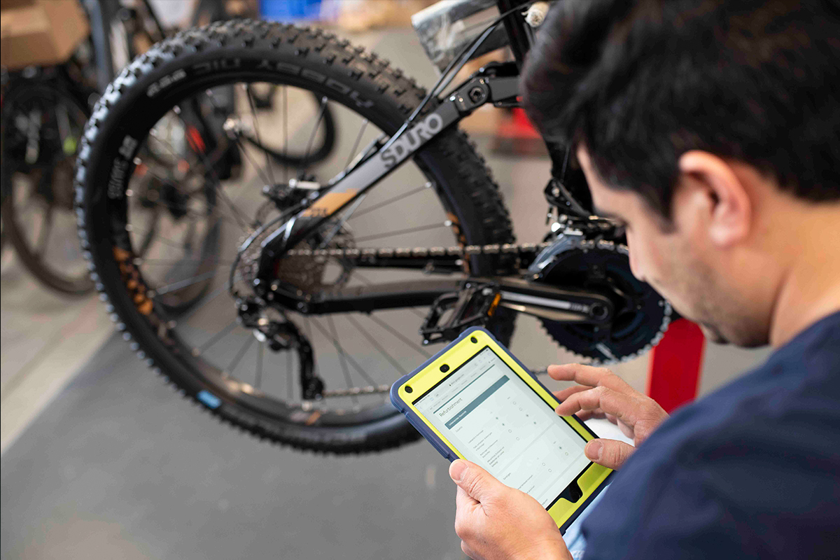 Detailaufnahme Mechniker mit Tablet, ließt Fahrraddaten aus, im Hintergrund Fahrrad im Anschnitt