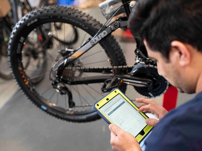 Detailaufnahme Mechniker mit Tablet, ließt Fahrraddaten aus, im Hintergrund Fahrrad im Anschnitt