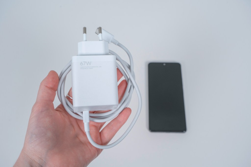 Hand hält weißes Ladekabel, im Hintergrund dunkles Smartphone auf weißem Tisch