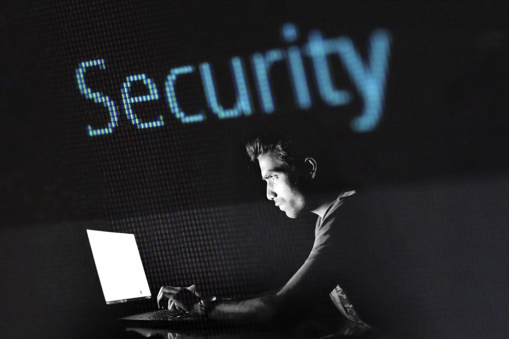 In einem schwarzen Raum sitzt ein Mensch vor einem geöffneten Laptop, der ihn anstrahlt. Darüber steht ein blauer Schriftzug "Security".