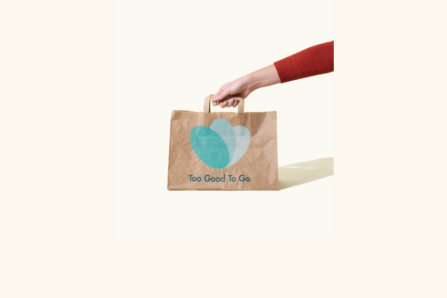 Auf einem beigen Untergrund greift eine Hand mit rotem Ärmel nach einer Papiertüte mit gründen Tot-Good-To-Go-Logo.
