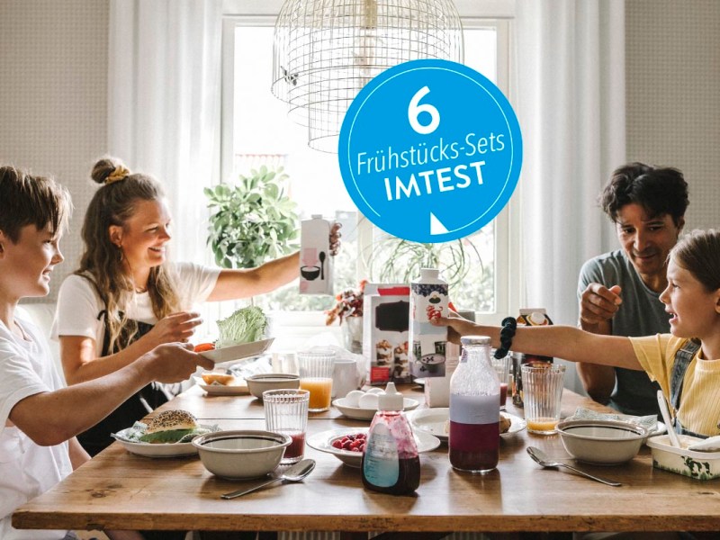 Vierköpfige Familie von der Seite am gedeckten Frühstückstisch mit blauem Button 6 Frühstückssets IMTEST