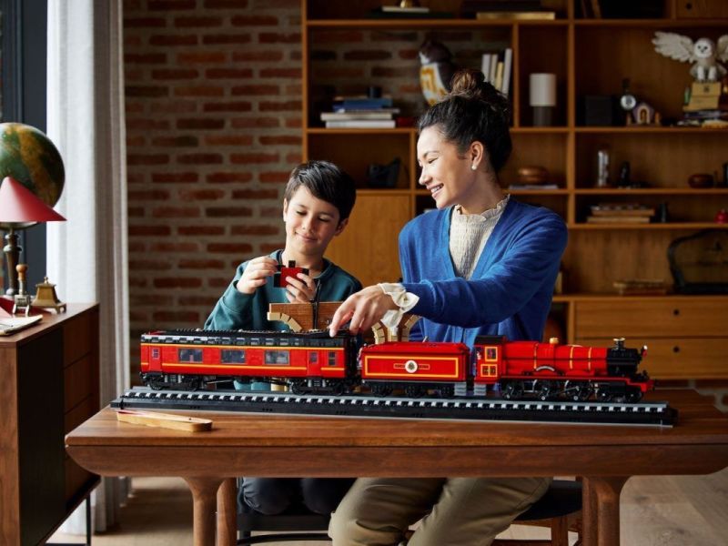 Frau und Junge hinter Tisch auf dem langer roter Zug aus Lego steht in Zimmer mit Holzregalen