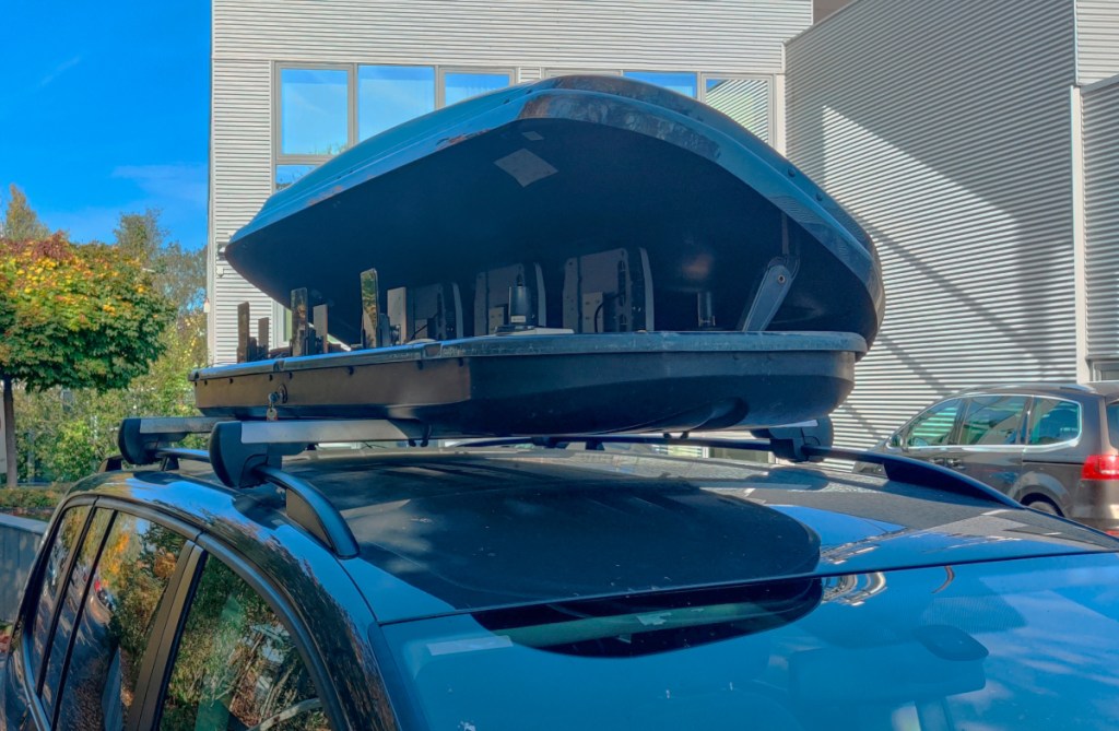 Geöffnete dunkle Dachbox auf dunkles Autodach vor weißem Gebäude
