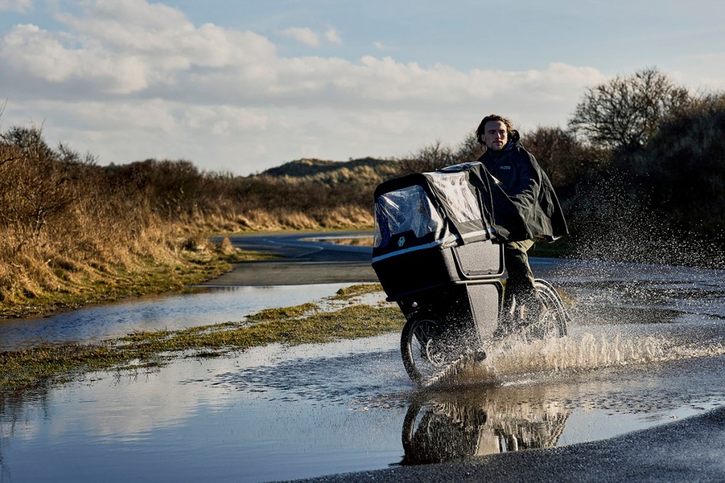 Mann fährt mit E-Cargo-Bike durch eine Pfütze in Dünenlandschaft bei herbstlichem Wetter