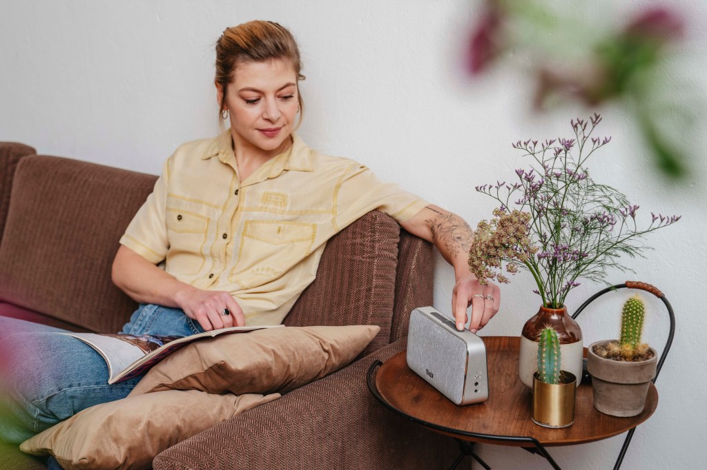 Eine Frau sitzt auf einem Sofa, von wo aus sie den Bluetooth-Speaker von Teufel bedient.