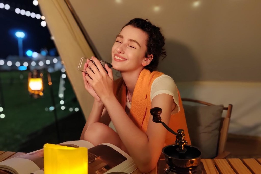 Eine brüntte junge Frau sitzt mit einer Tasse Kaffee in einem Zelt und lächelt.