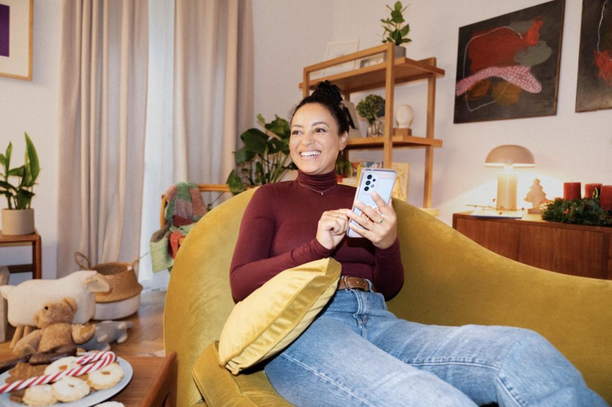 Eine Frau sitzt auf einem gelben Sofa mit einem Smartphone in der Hand und lacht