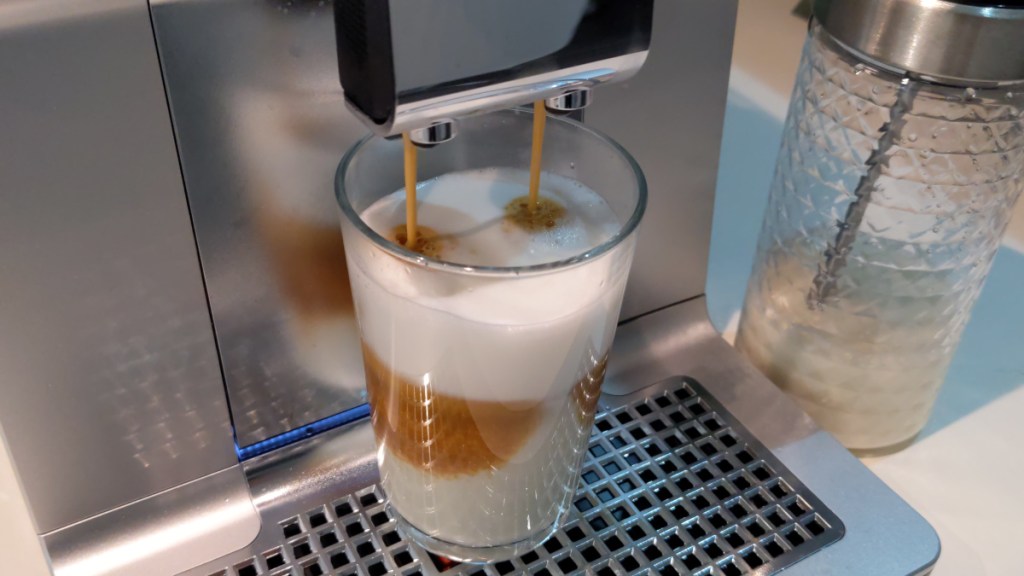 Glas unter silbernem Kaffeevollautomaten aus dem Latte Macchiato rausläuft