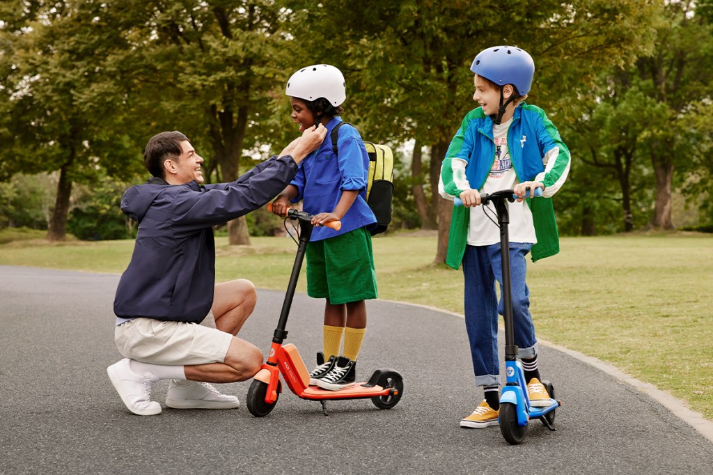 zwei Kinder auf je einem KQi Youth Kick-Scooter im Park, daneben ein Erwachsener, der den Sitz des Helms bei einem Kind prüft