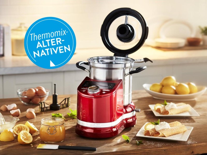 Eine Küchenmaschine von KitchenAid auf einem Holztisch neben Lebensmitteln