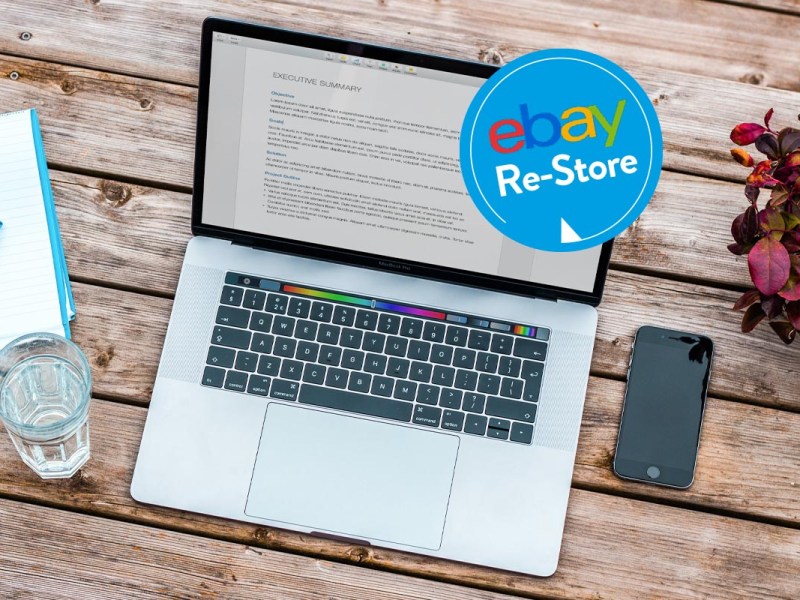 Das MacBook Pro auf einem Holztisch. Rechts daneben ein iPhone-Modell und lins eine Glas gefüllt mit Wasser. An der Bildschirmecke rechts oben ein blauer Kreis mit dem eBay-Logo und ein weißer Schriftzug in den Worten Re-Store drunter.