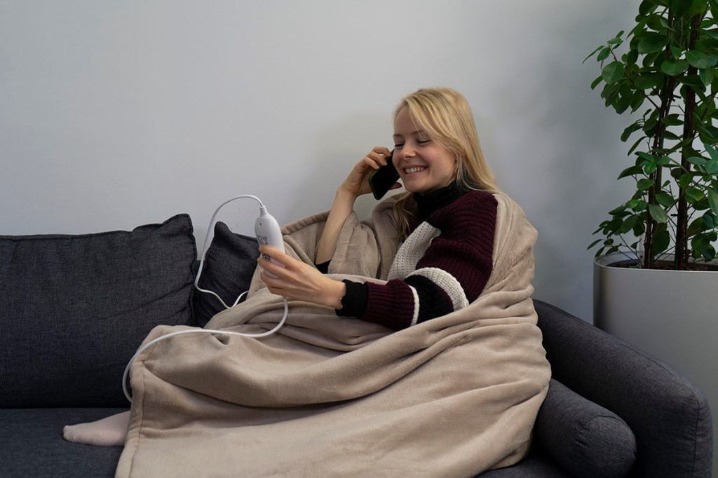 Frau in Heizdecke eingekuschelt sitzt auf einem Sofa und telefoniert mit einem Smartphone.