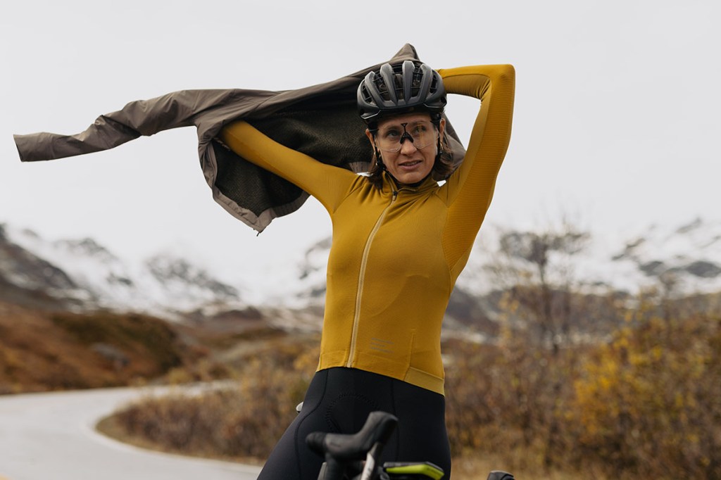 Frau auf Rennrad vor schneebedeckten Bergen, die sich eine wärmende Weste überzieht.