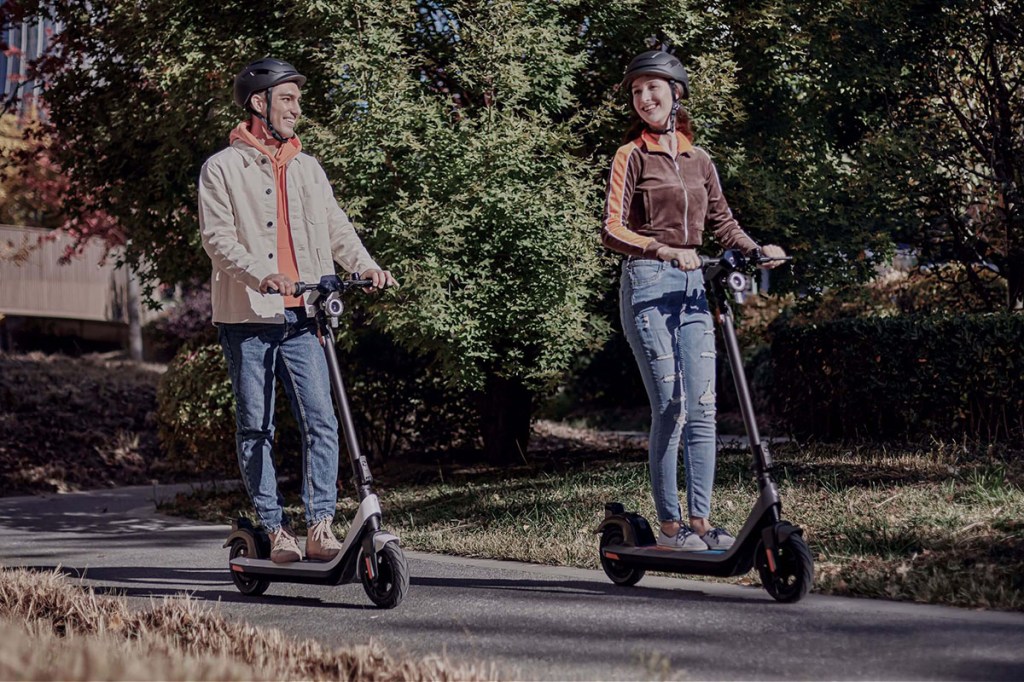 Frau und Mann fahren je auf einem E-Scooter durch Parklandschaft.