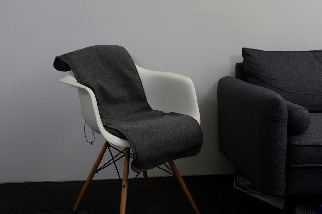 Eine graue Heizdecke liegt auf einem weißen Stuhl und bedeckt Sitzfläche sowie Rückenlehne. Der Stuhl steht neben einem dunkelgrauen Sofa rechts im Bild. Beide Möbelstücke befinden sich vor einer hellgrauen Wand