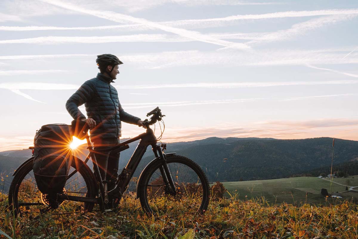 Mann mit seinem E-bike in der Natur auf einem Hügel, im Hintergrund die untergehende Sonne
