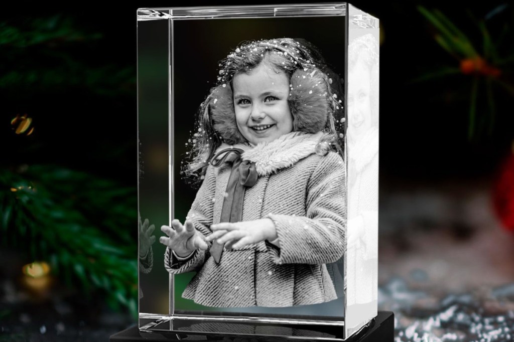 Rechteckiger hochkant gestellter durchsichtiger 3D-Glasklotz schräg von vorne mit Foto von Mädchen in Wintermantel und mit Ohrschützern, das lächelt vor dunklem Hintergrund mit Tannenzweigen links