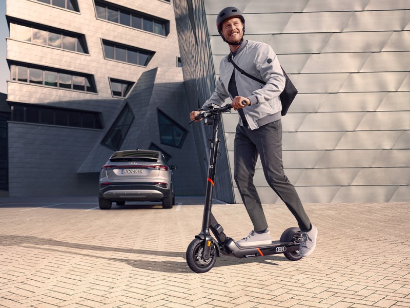 Mann fährt auf dem Audi electric kick scooter powered by Egret, im HIntergrund steht ein E-Auto von Audi