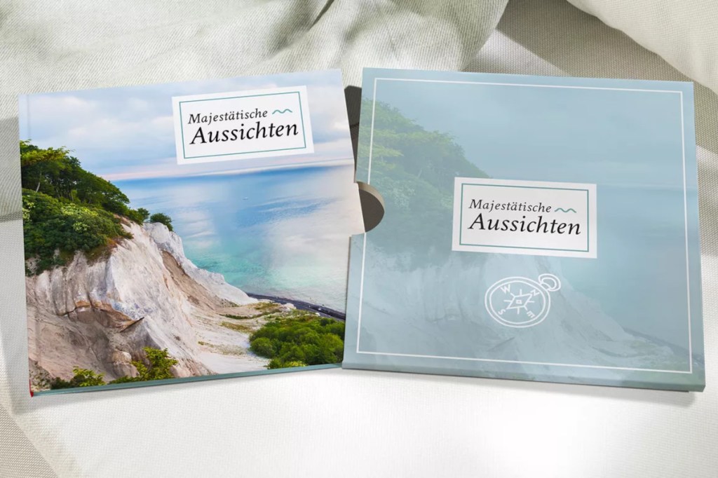 Auf hellen Leinentüchern  links ein Fotobuch, das eine weiße Steilküste am blauen Meer zeigt und rechts ein blauer Schuber mit der Aufschrift "Wunderschöne Aussichten"
