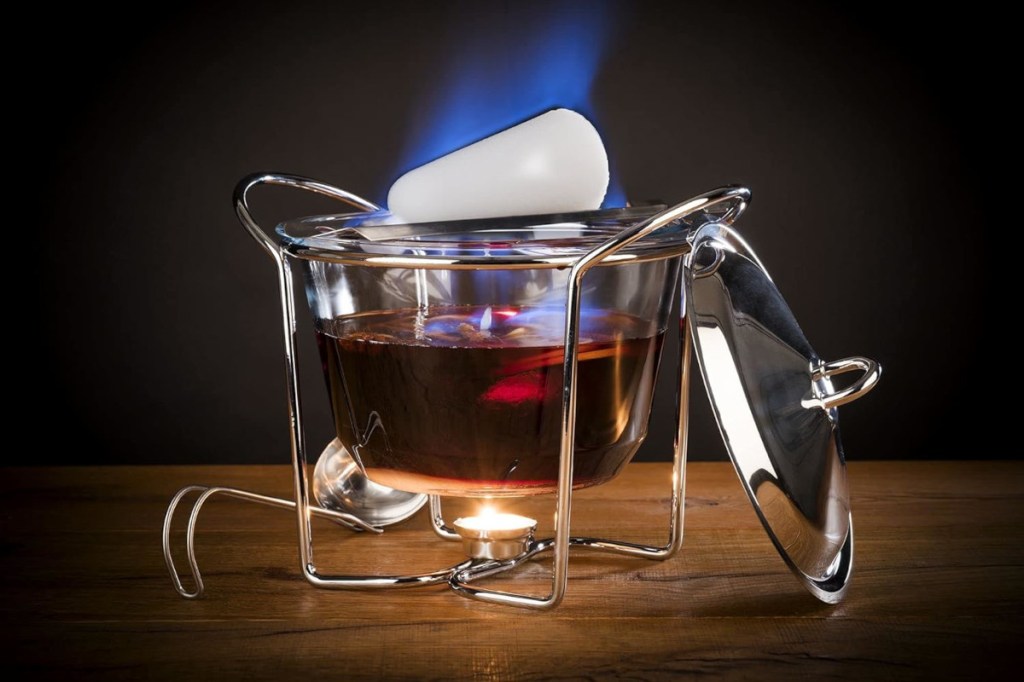 Metallgestell mit Glasbehälter, der mit rotem Getränk gefüllt ist und über Flamme von Teelicht hängt, ein weißer Zuckerhut oben drauf, ein silberner Deckel angelehnt daneben auf Holzfläche vor dunklem Hintergrund