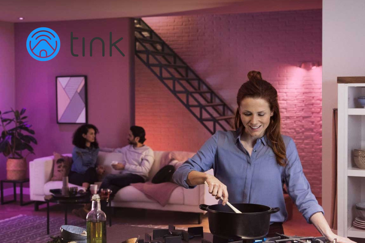 Frau kocht im Vordergrund, im Hintergrund Mann und Frau auf Sofa, pinkes Licht, oben links Tink Logo
