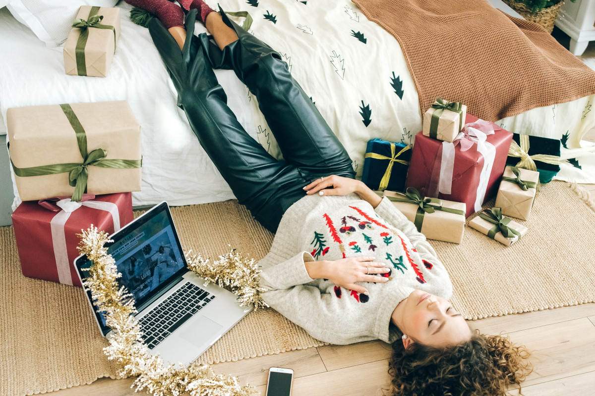 Junge Frau liegt auf Boden mit Beinen auf Bett zwischen Weihnachtsgeschenken und neben aufgeklapptem Laptop