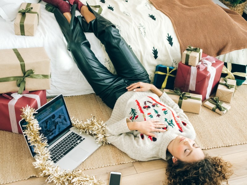 Junge Frau liegt auf Boden mit Beinen auf Bett zwischen Weihnachtsgeschenken und neben aufgeklapptem Laptop