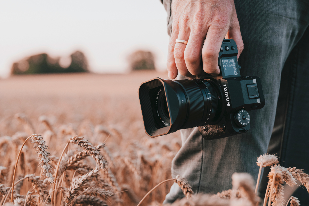 Schwarze Fujifilm X-H2 Kamera wird von Hand an Bein gehalten, Person steht im Getreidefeld