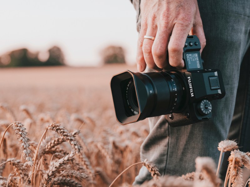 Schwarze Fujifilm X-H2 Kamera wird von Hand an Bein gehalten, Person steht im Getreidefeld