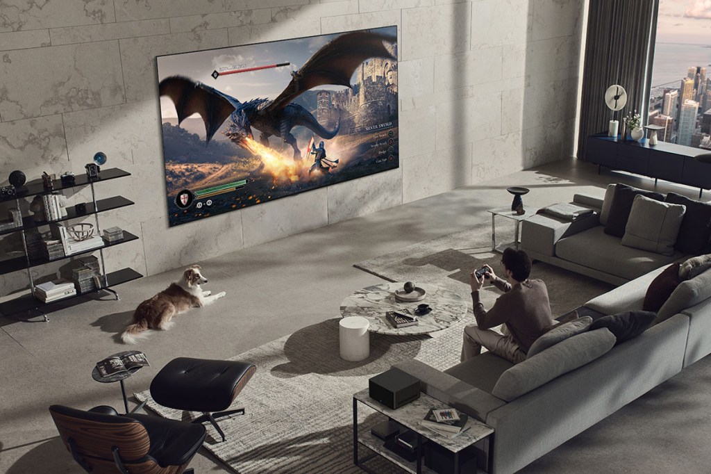 Mann sitzt im Wohnzimmer vor einem XXL-OLED-TV von LG (LG SIGNATURE OLED TV M3), der flach an einer Wand hängt.