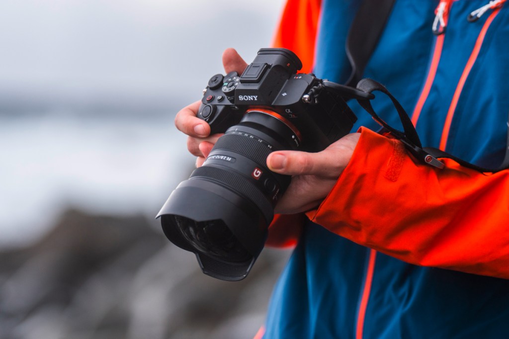 Hände von Mensch in bunter Regenjacke halten schwarze Sony Alpha 7R V Kamera nach unten gerichtet vor sich