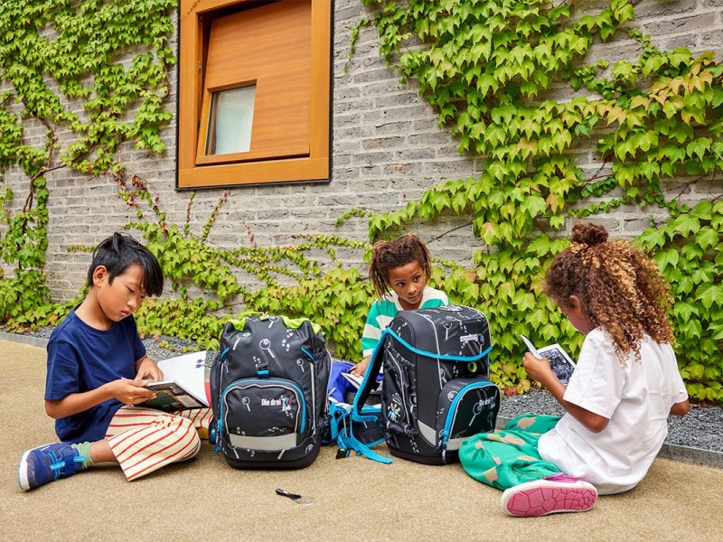 Drei Kinder sitzen entspannt mit ihren Ranzen vor der Schule