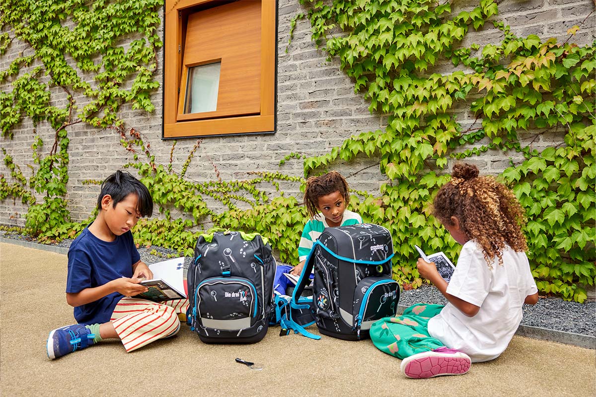 Drei Kinder sitzen entspannt mit ihren Ranzen vor der Schule