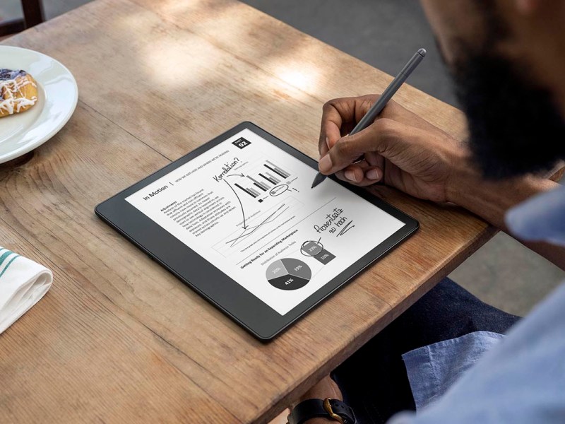 Mann schreibt auf eingeschaltetem Amazon Kindle Scribe auf hellem Holztisch