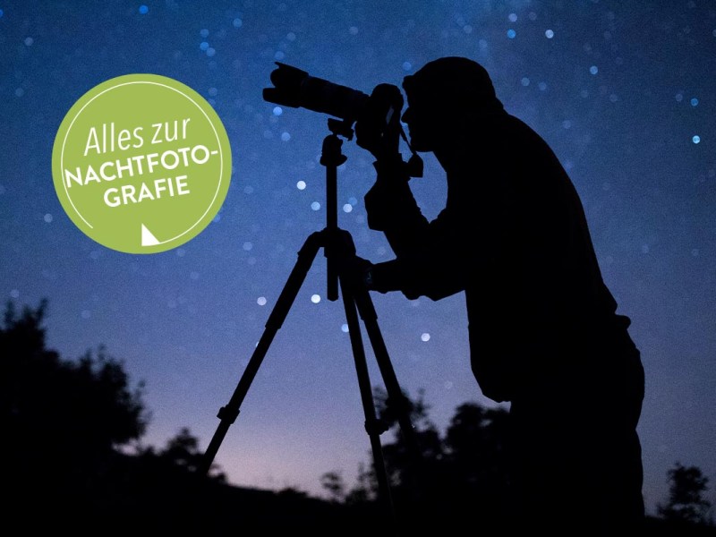 Eine Nachtbildaufnahme einer Person, die mit einem Stativ den Nachthimmel fotografiert. Im Hintergrund ist der Nachthimmel mit Sternen in dunkelbla