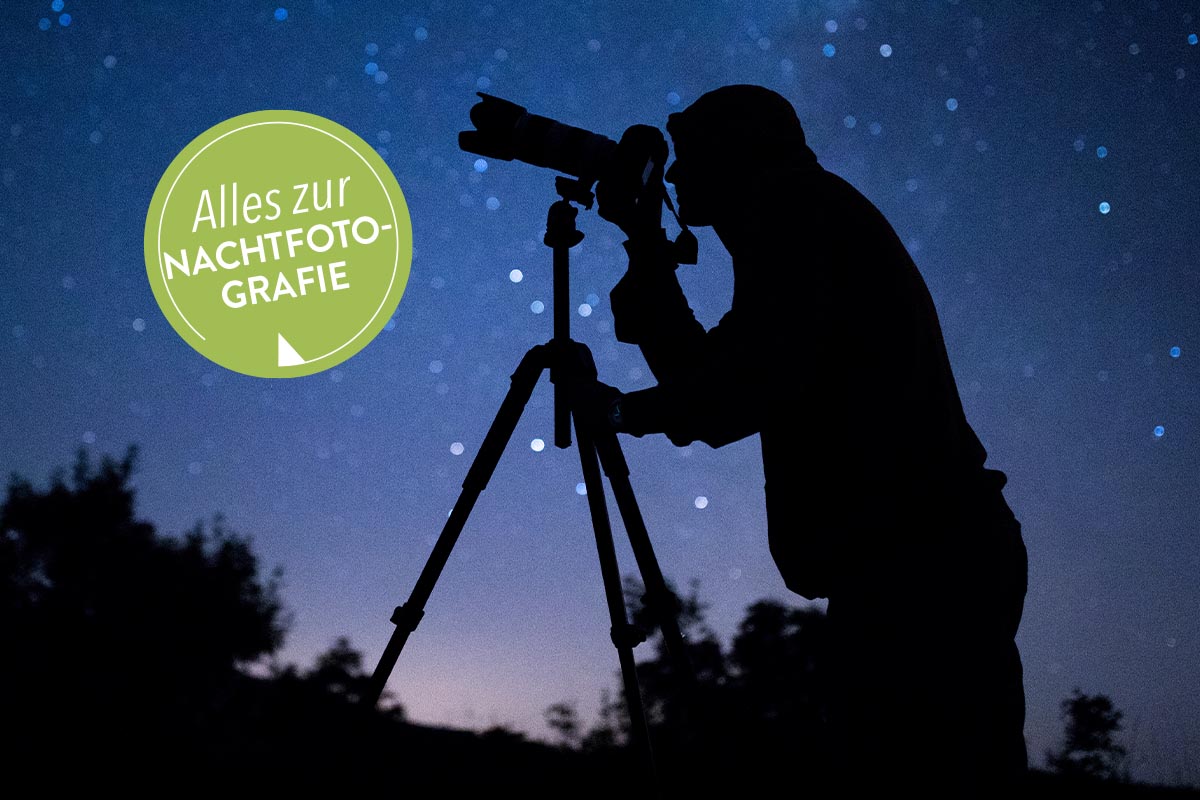 Eine Nachtbildaufnahme einer Person, die mit einem Stativ den Nachthimmel fotografiert. Im Hintergrund ist der Nachthimmel mit Sternen in dunkelbla
