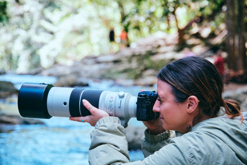 Frau an Fluss hält Kamera mit großem weißen Teleobjektiv vor Auge von der Seite
