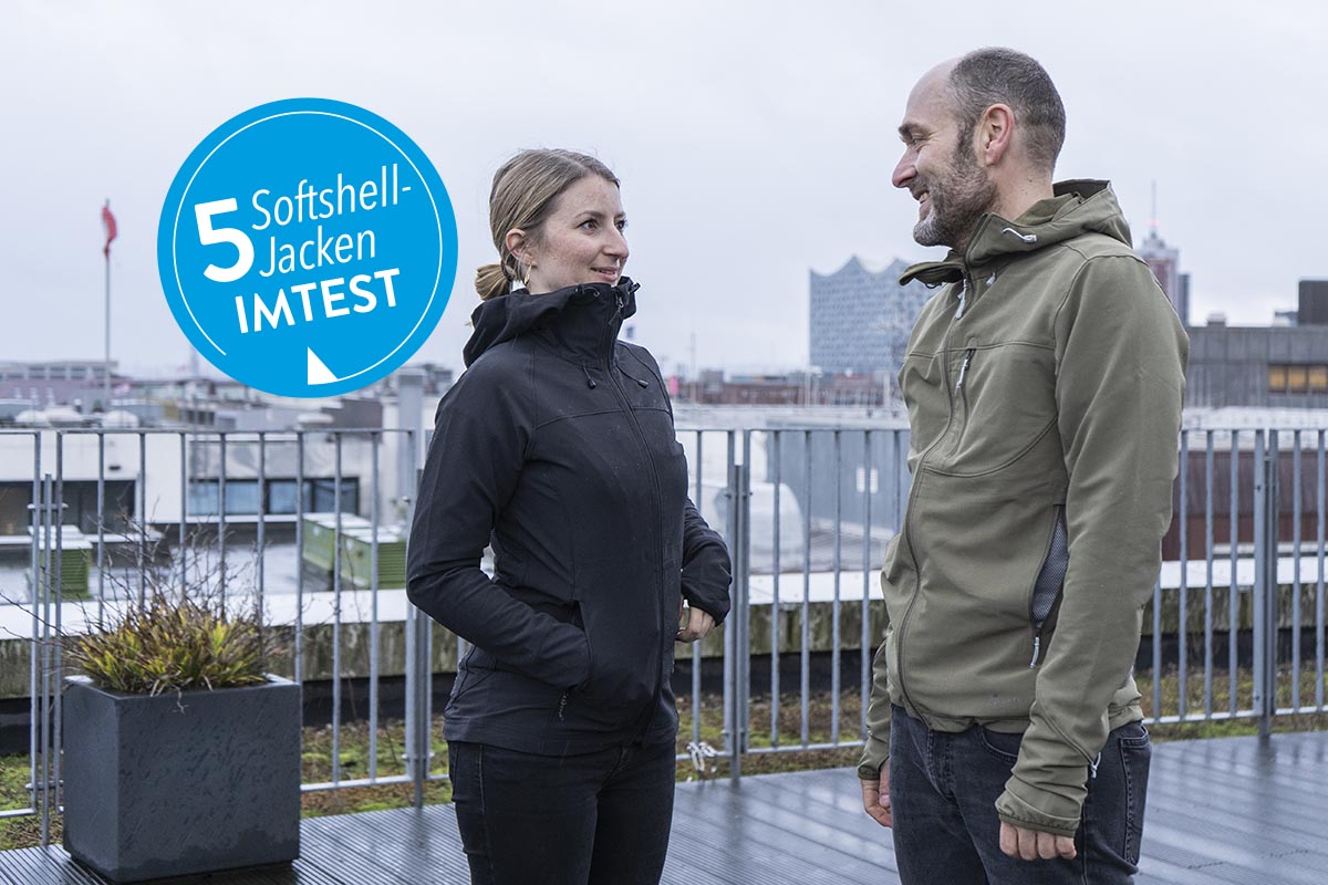 Eine Frau und ein Mann, bekleidet u.a. mit einer Softshell-Jacke stehen auf einer Dachterrasse mit Blick auf eine Stadt-Skyline.