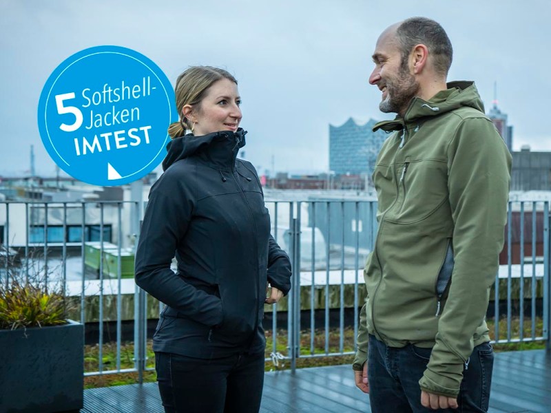 Eine Frau und ein Mann, bekleidet u.a. mit einer Softshell-Jacke stehen auf einer Dachterrasse mit Blick auf eine Stadt-Skyline.