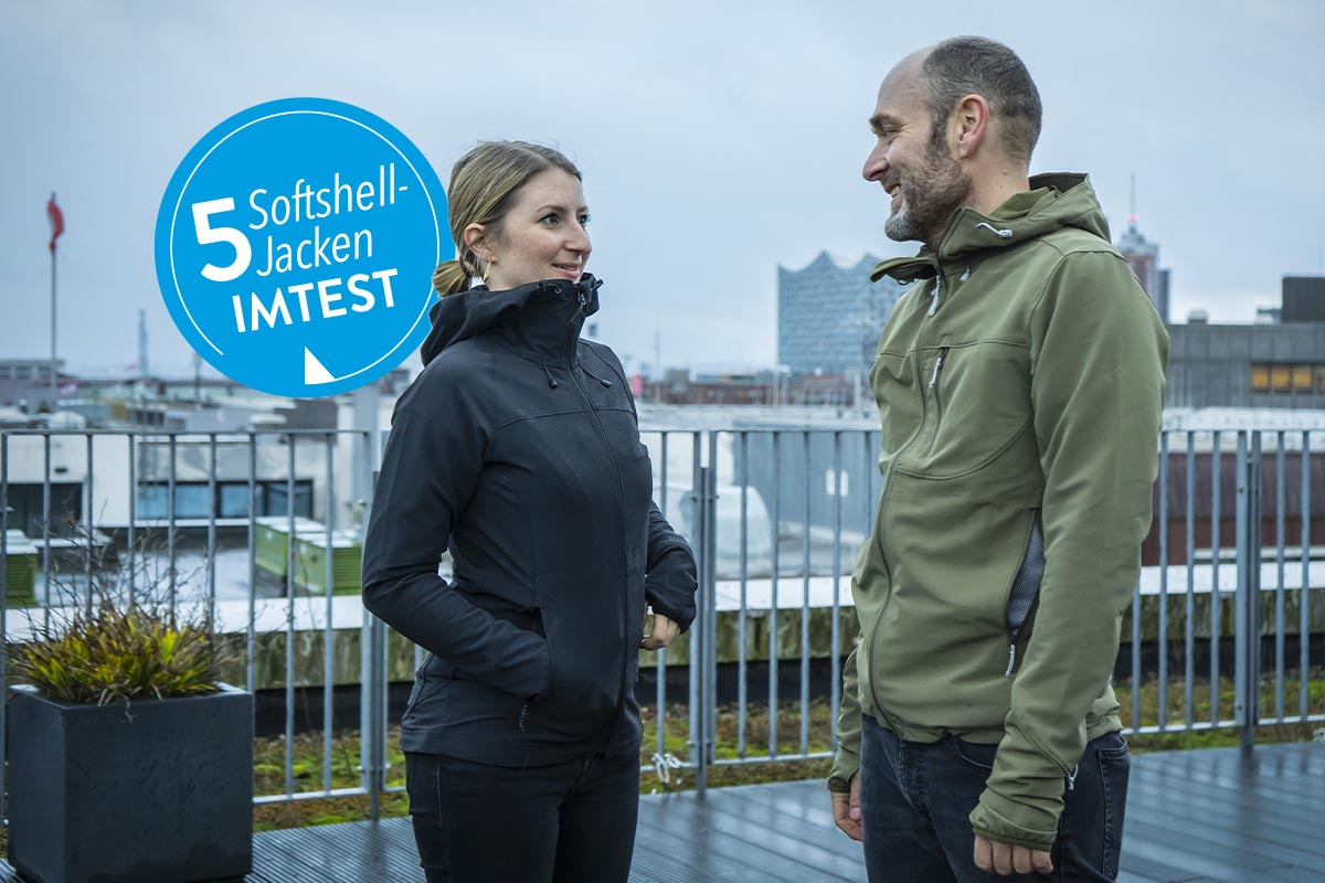 Eine Frau und ein Mann, bekleidet u.a. mit einer Softshell-Jacke stehen auf einer Dachterrasse mit Blick auf eine Stadt-Skyline.