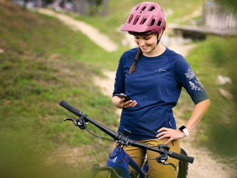 Frau sitzt auf ihrem Mountainbike, im Hintergrund ein Schotterweg im Grünen, sie guckt auf ihr Smartphone