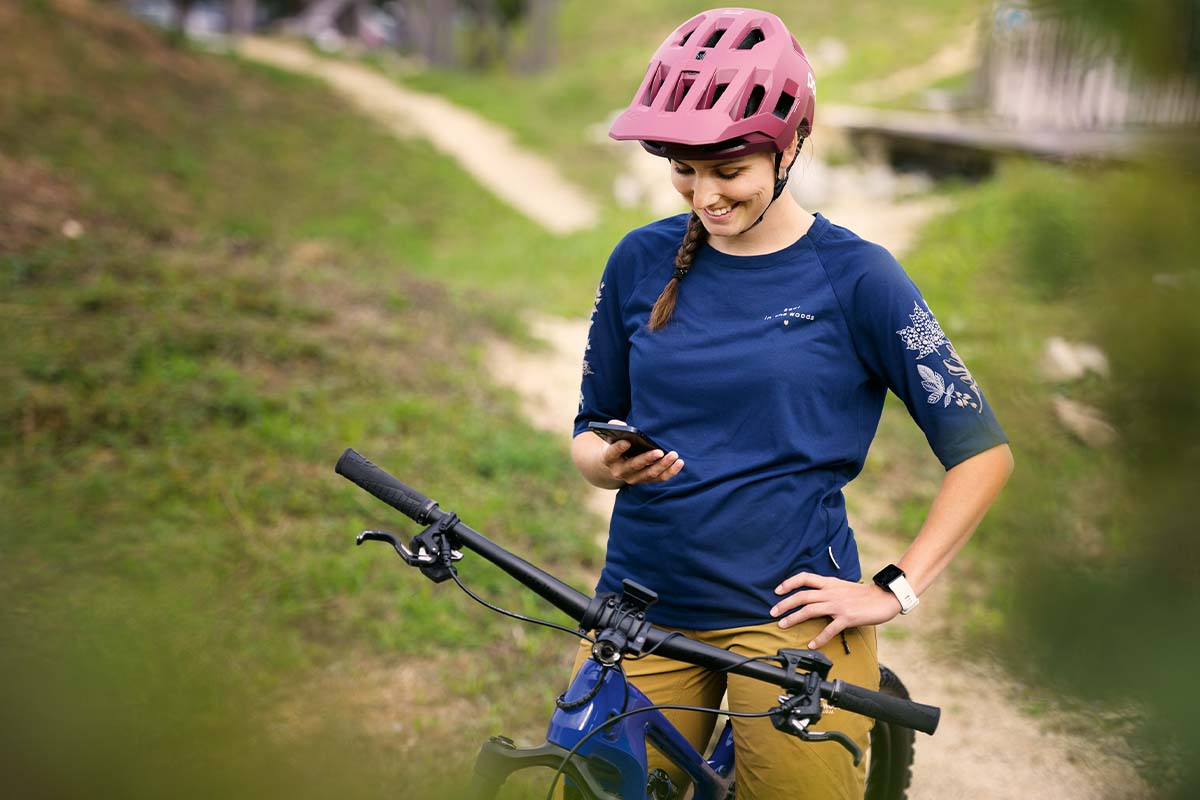 Frau sitzt auf ihrem Mountainbike, im Hintergrund ein Schotterweg im Grünen, sie guckt auf ihr Smartphone