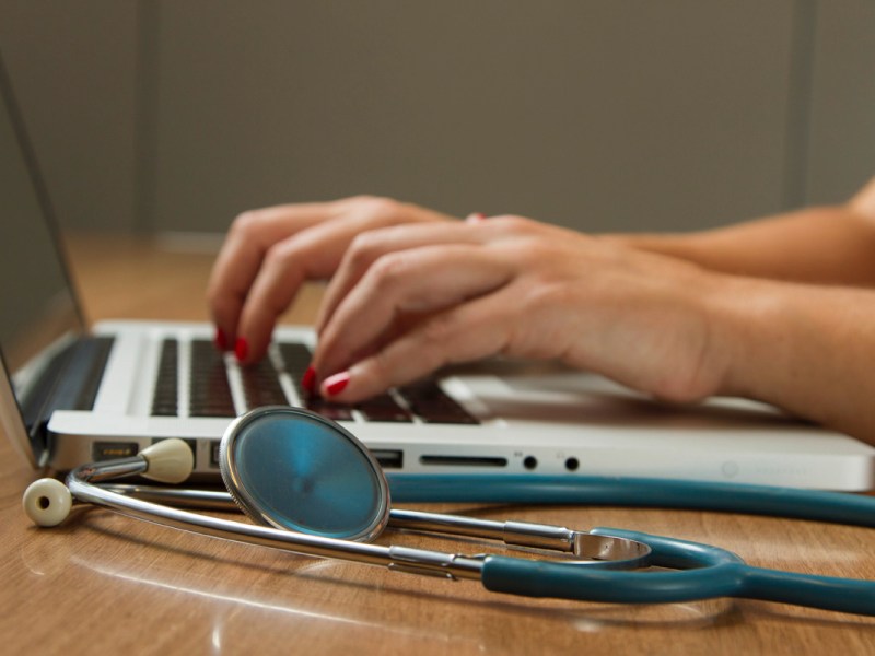 Eine Frau tippt auf einem Laptop, neben ihr liegt ein Stetoskop.