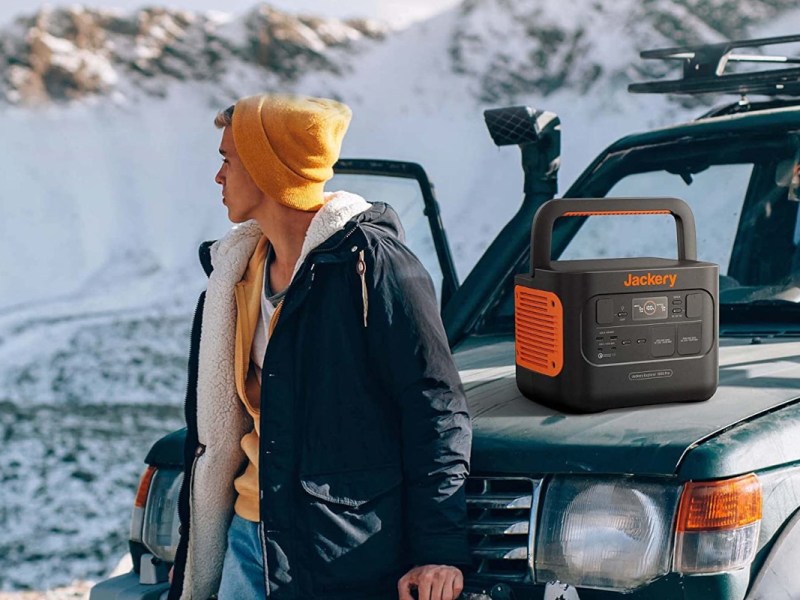 Junger Mann in Winterkleidung lehnt an Offroad-Auto auf dessen Motorhaube eine Jackery Power Station steht in Schneelandschaft
